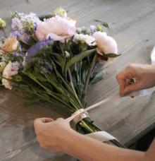 En person binder ett band runt en bukett av färgglada blommor på ett träbord.