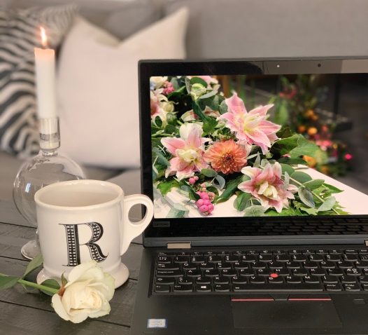 En bärbar dator med en bild av blommor på skärmen står på ett bord bredvid en tänd vit stearinljus och en vit kopp med bokstaven "R", samt en vit ros. I bakgrunden syns en grå soffa med kuddar.